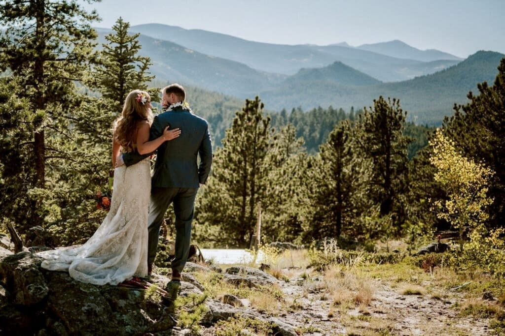 A bride and groom stand arm-in-arm looking out at forest and mountains