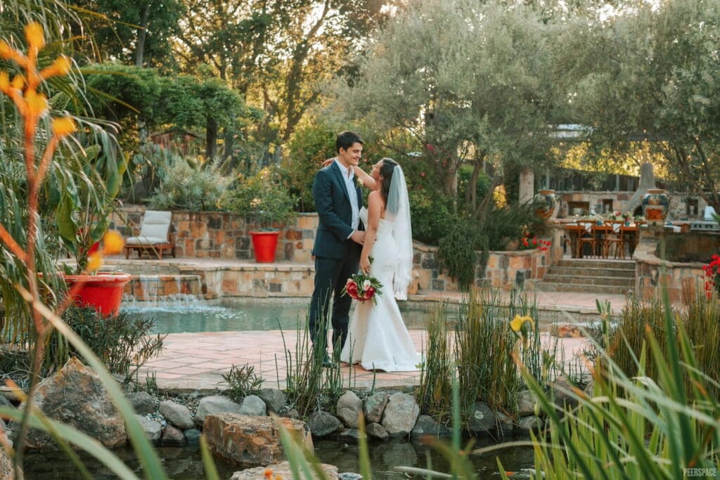 A bride and groom embrace by a pool with a small, casual wedding reception set up in the background