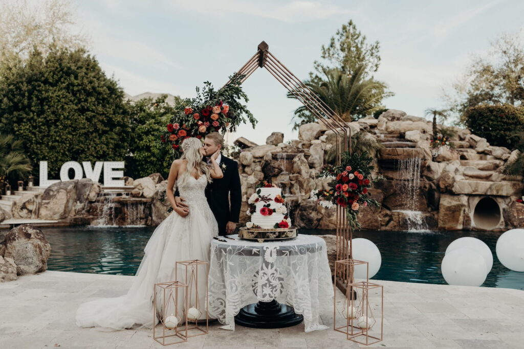 A bride and groom kiss while standing next to their wedding cake at an outdoor venue with a beautiful pool with rock formations and waterfalls in the background
