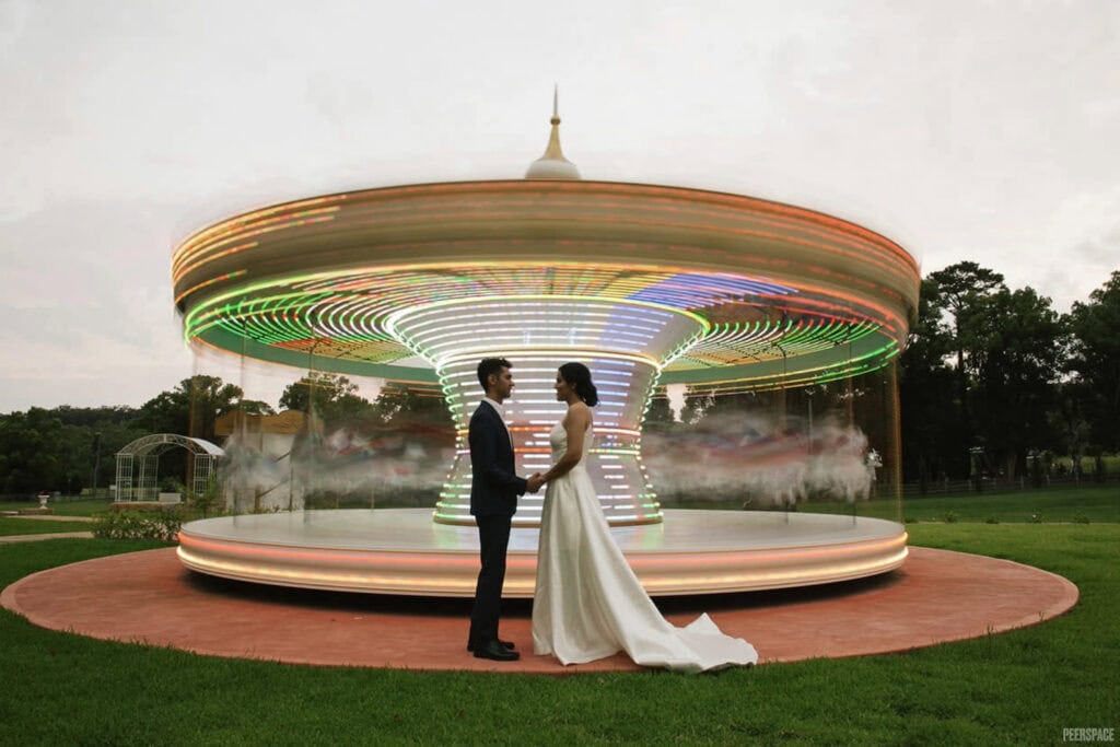 A bride and groom stand in front of a carousel in motion