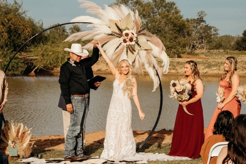 A casually-dressed groom raises his and his bride's hand as she smiles at their wedding guests while standing at an outdoor altar made of a round iron ring decorated in feathers and flowers