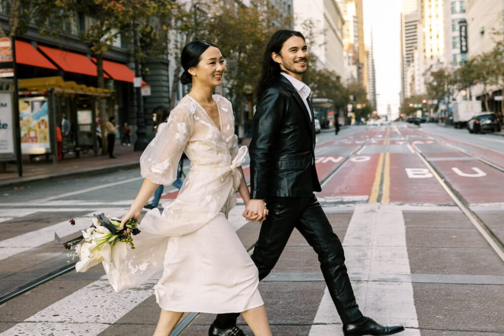 A bride and groom cross the street hand in hand in the daytime