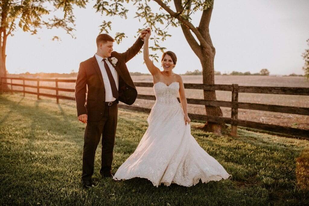 A groom spins his bride in a meadow by a wooden fence with the sun setting behind them