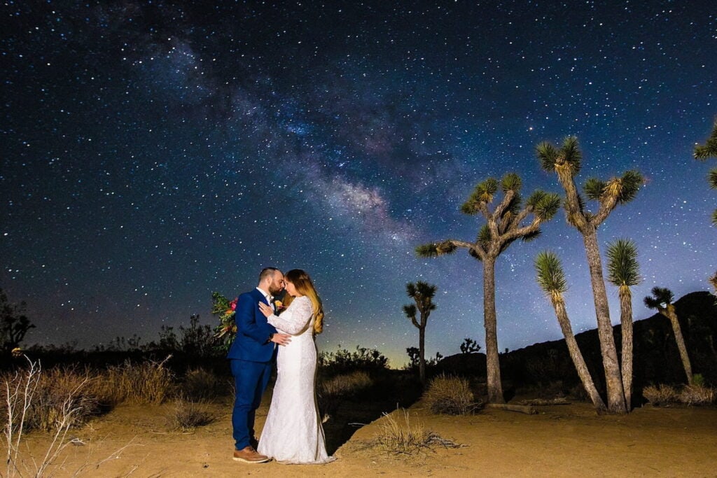A bride and groom stand outside in the desert under the night sky, which is illuminated by a variety of stars