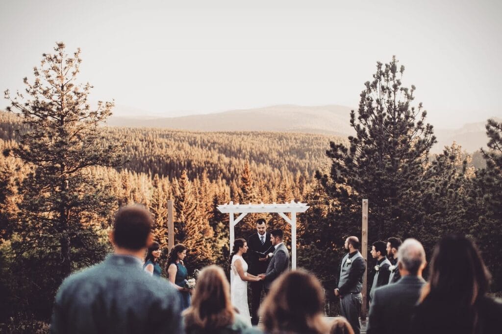 A bride and groom exchange vows under a white wooden pergola at the top of a hill overlooking forest and mountains