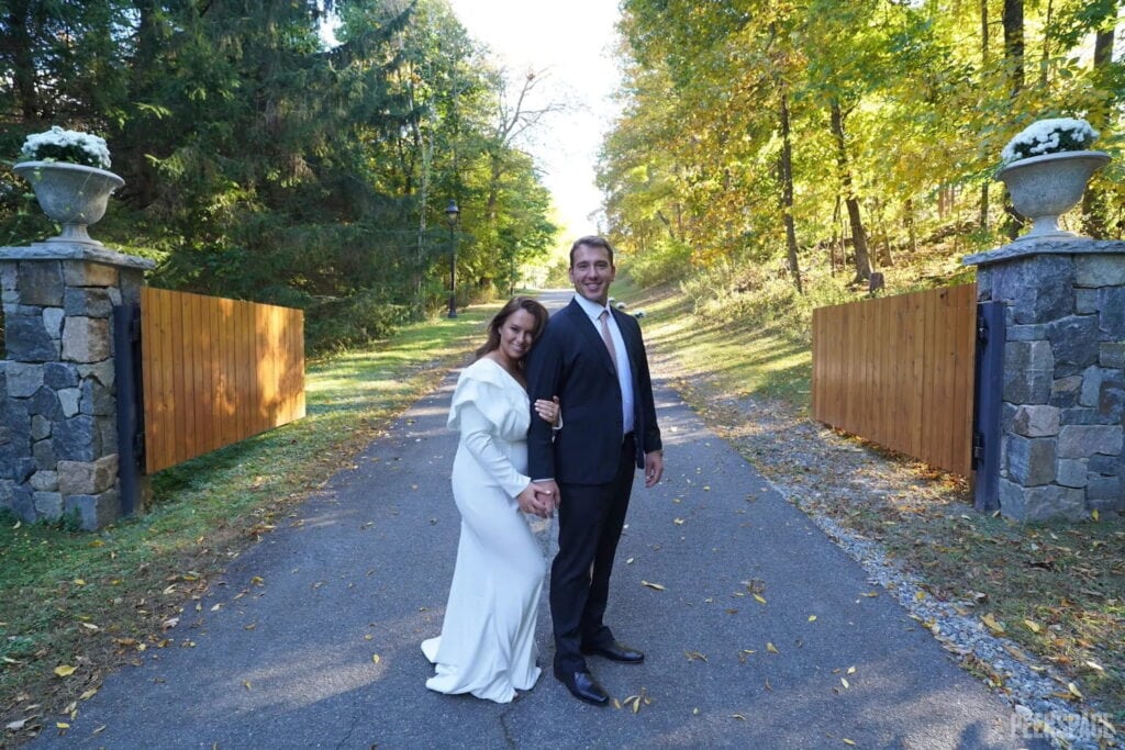 A bride and groom pose at an entry gate of a wedding venue in a rustic location