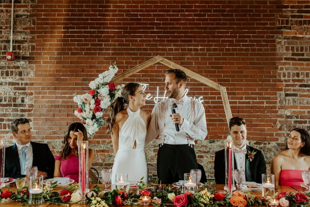 A bride and groom stand at their head table surrounded by their bridal party and give a speech while in front of a brick wall and a wooden arch decorated with flowers