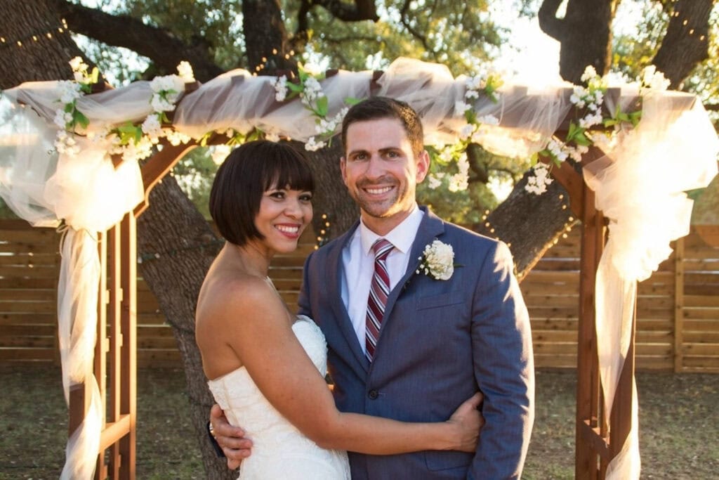 A bride and groom smile under a wooden canopy decorated in garland