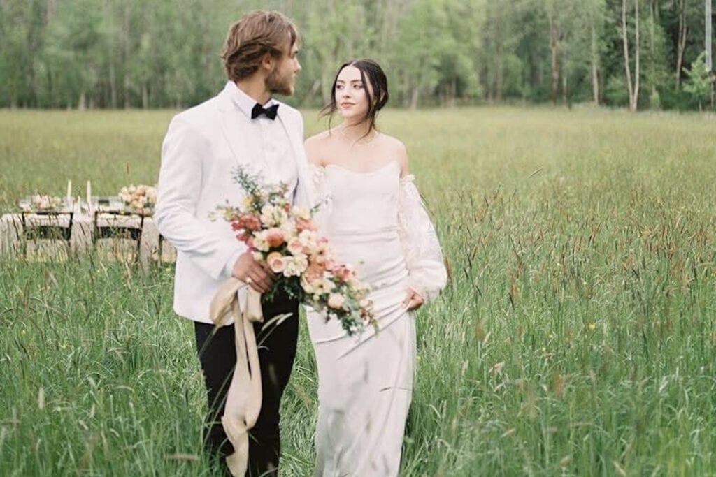 A bride and groom walk in a meadow with a boho-styled reception table in the background