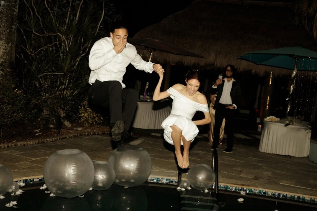 A bride and groom gleefully jump in a pool dressed in their wedding clothes