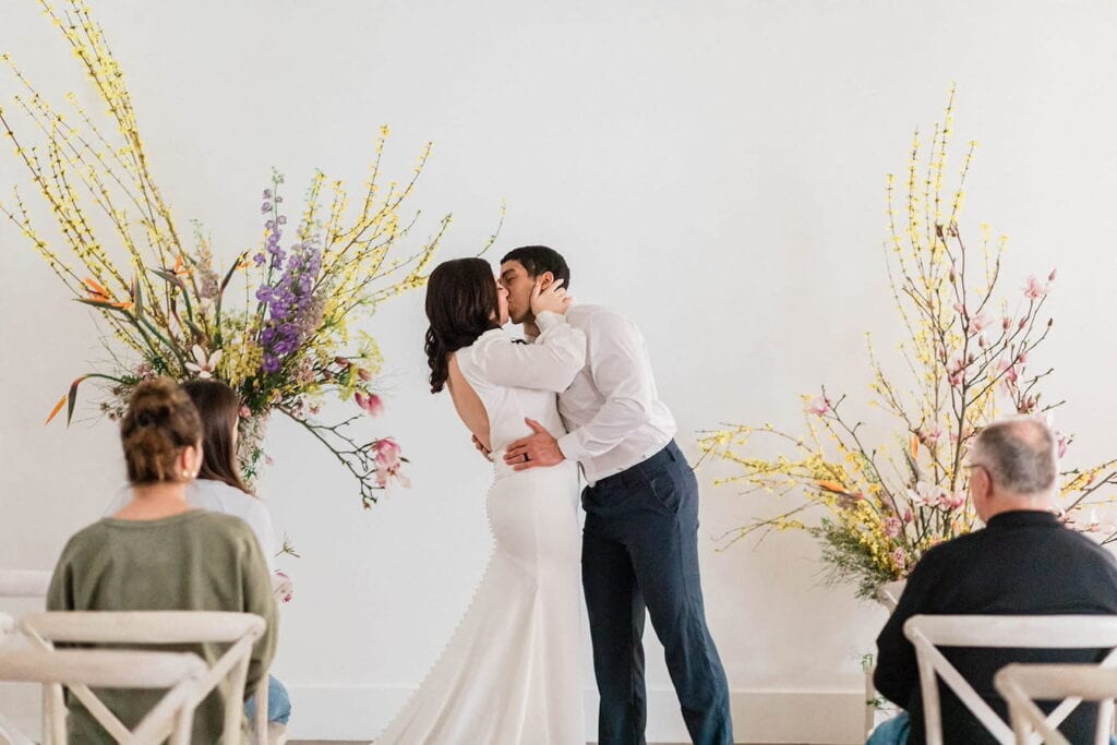 A bride and groom kiss in front of friends and family at a simply decorated altar in a bright, white event space