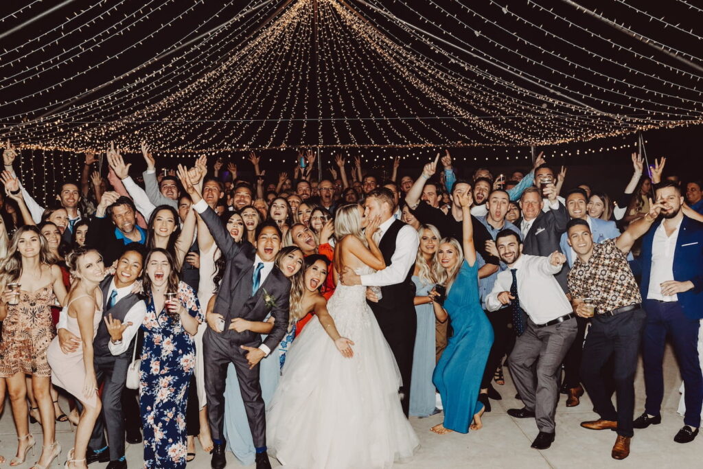 A bride and groom kiss in a sea of friends and family who are surrounding them, cheering while posing for the camera