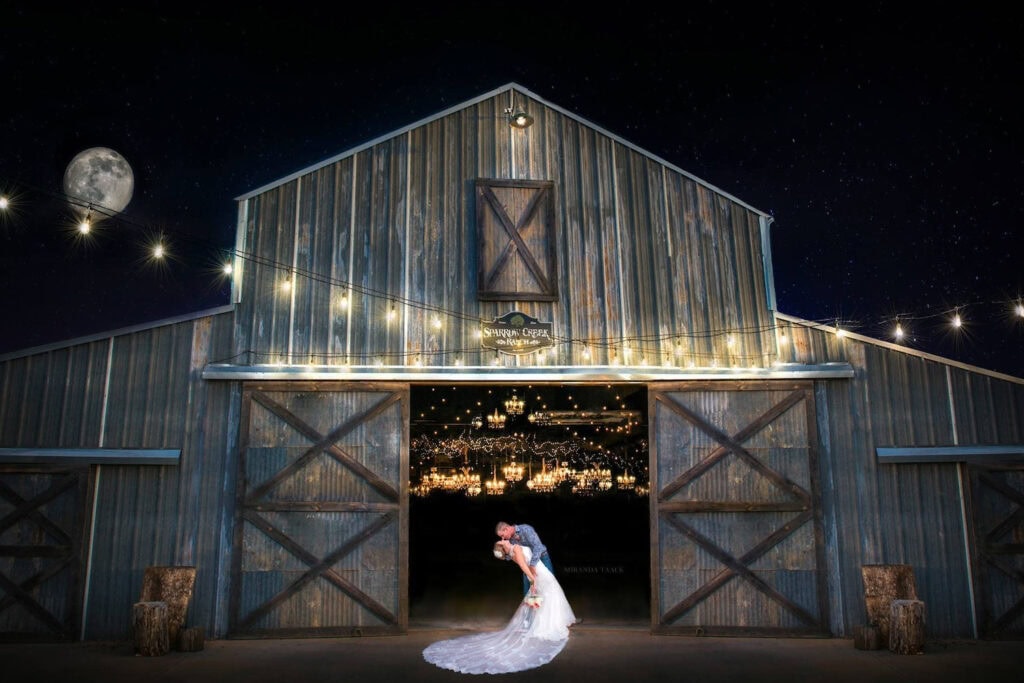 A bride and groom kiss outside of a huge barn at night under string lights with a huge full moon seen in the distance