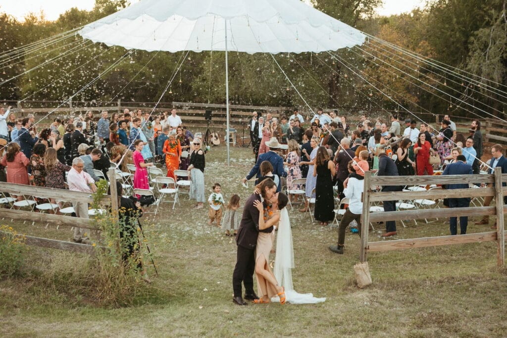A newly-married couple kisses at the end of an aisle set up outside in a horse pen