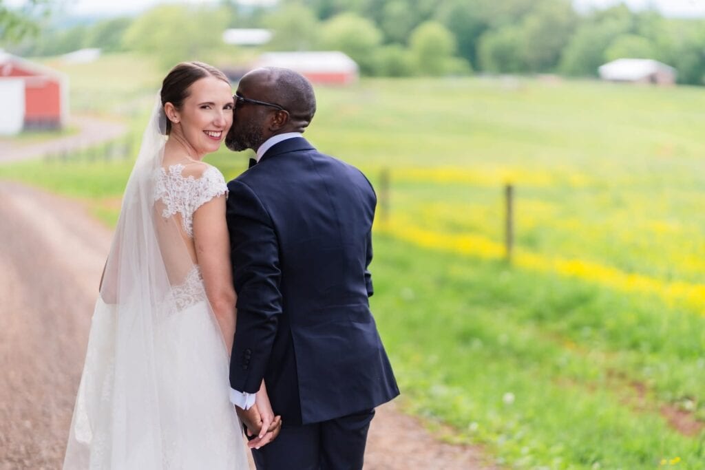 A groom kisses his bride's cheek as she looks back at the camera as they stand on a dirt path by a red barn