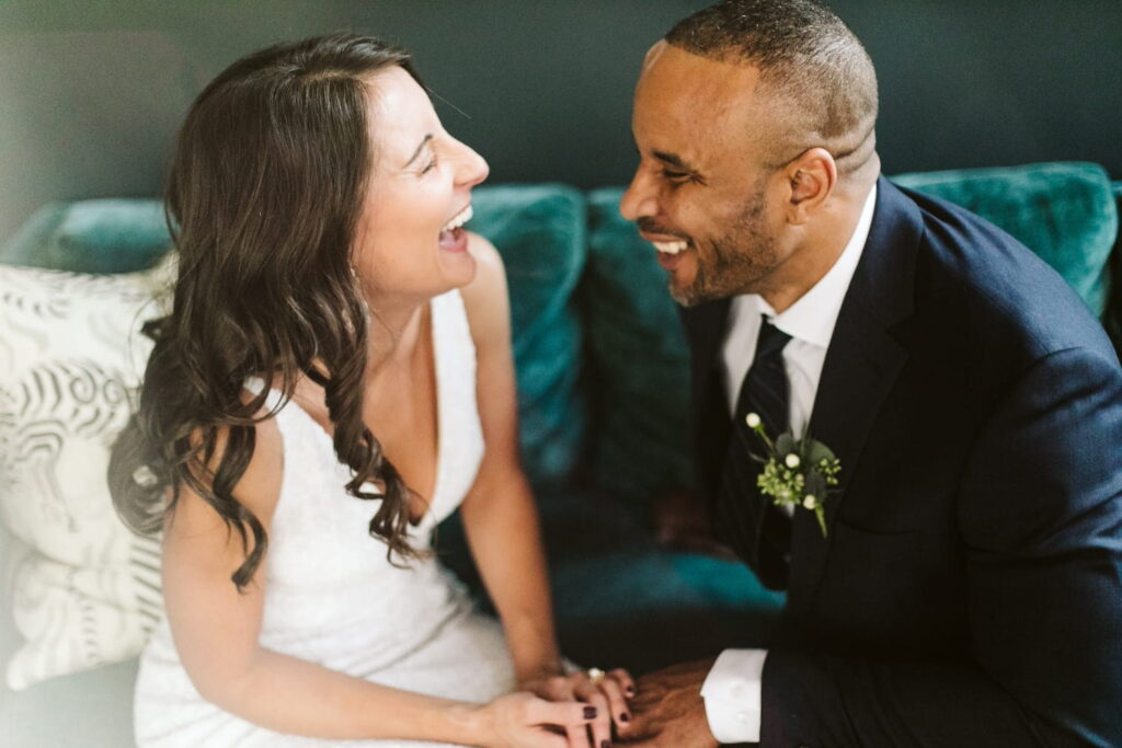 A bride and groom share a laugh while sitting on a couch