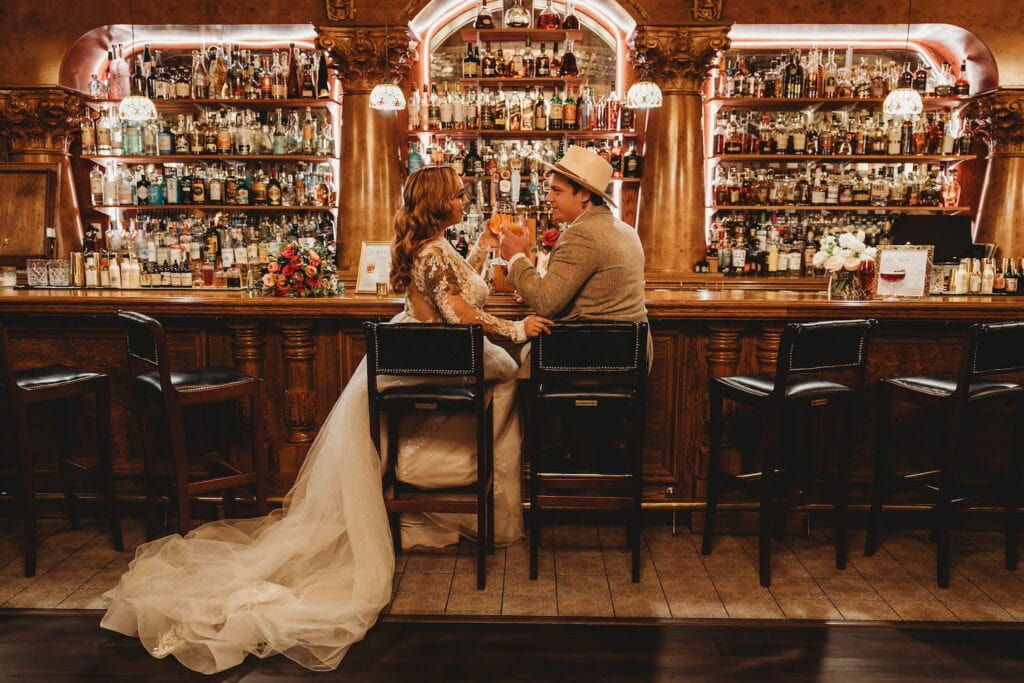 A bride and groom sit on barstools at an old-timey looking bar toasting one another