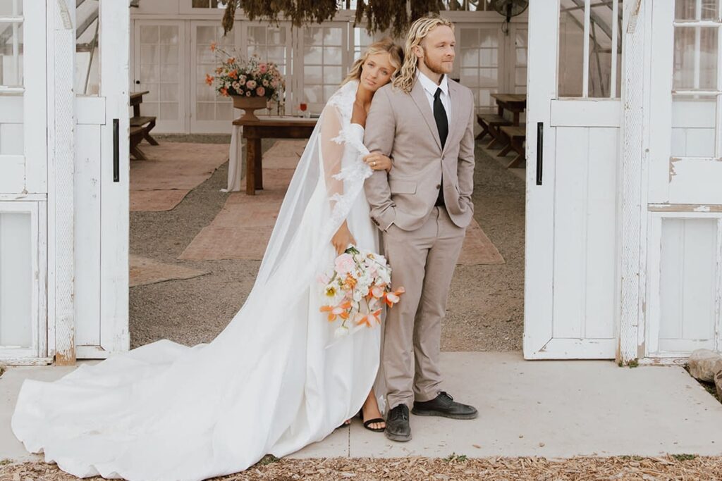 A bride and groom steal a quiet moment outside of an all-white barn with the sliding doors open to reveal brightly-colored floral arrangements