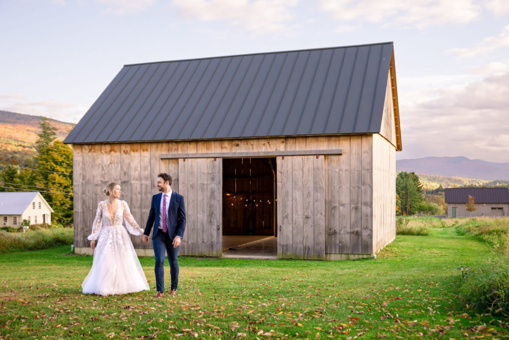 Bride and groom walk hand in hand with small barn behind them and vast farmland beyond