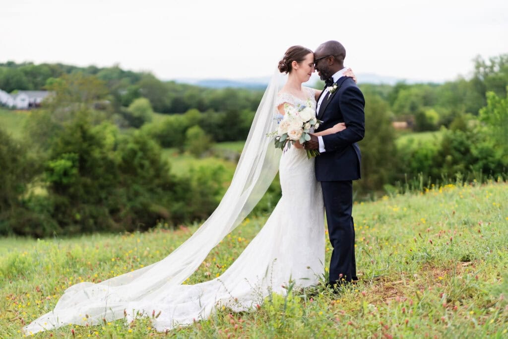 A bride and groom share a tender moment in a field on a hill with a house in the distance