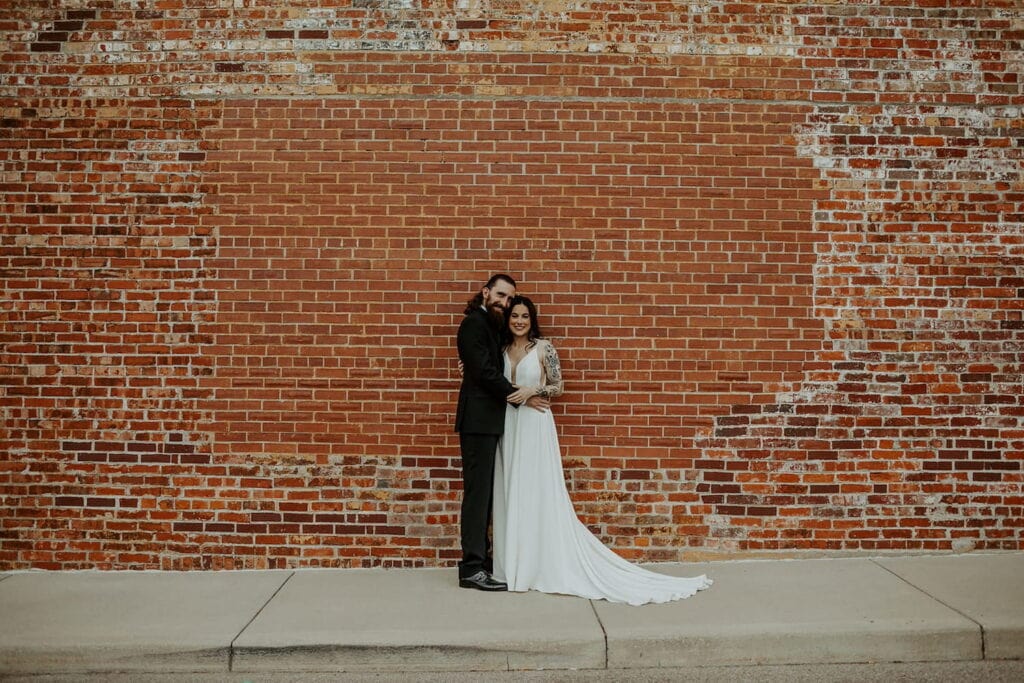 A bride and groom pose in front of a large brick wall