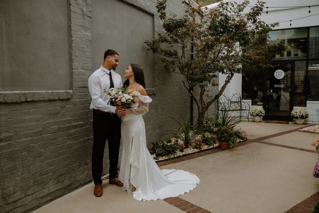 A bride and groom pose by a gray brick wall looking intently into each other's eyes