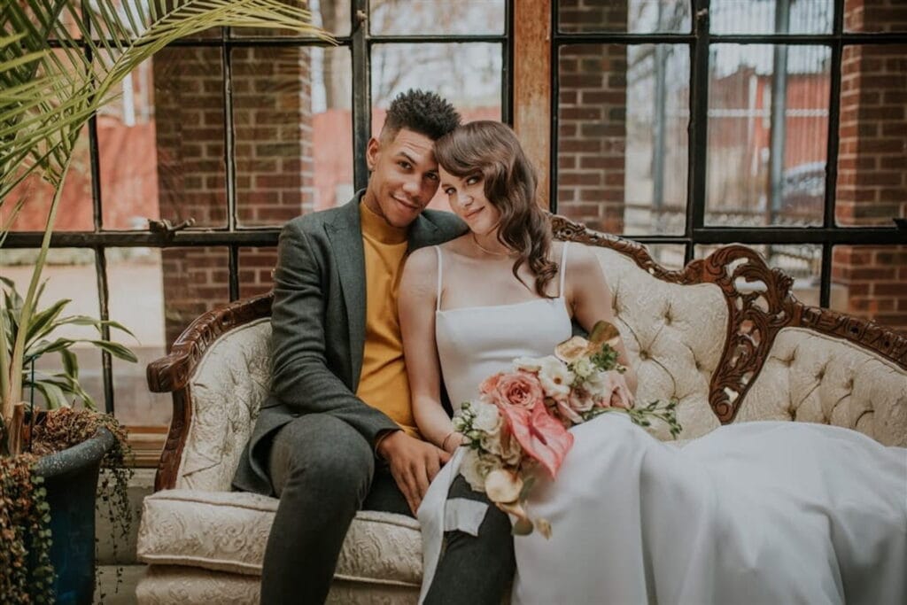 A bride and groom pose on a chaise surrounded by windows looking out onto a courtyard