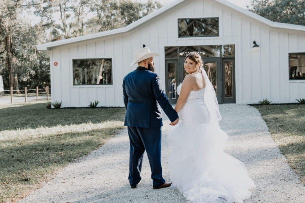 A bride and groom hold hands while walking towards a small, rustic barn building