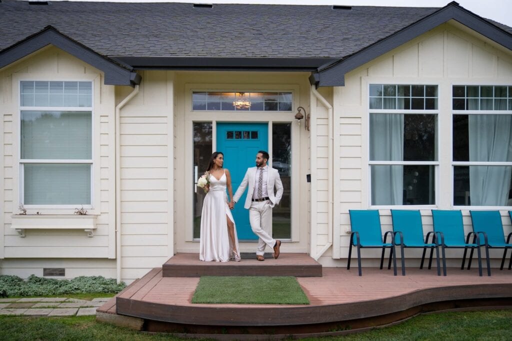 A bride and groom stand in front of an aqua door with matching aqua chairs to the right