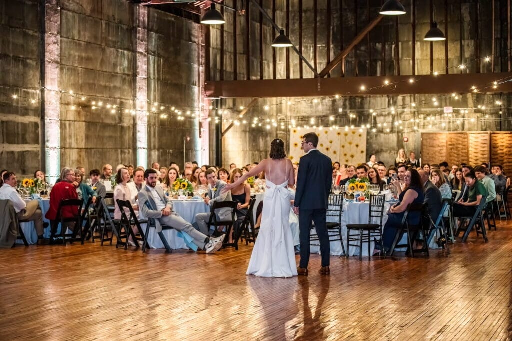 A bride and groom address their guests under twinkling string lights in an industrial reception venue
