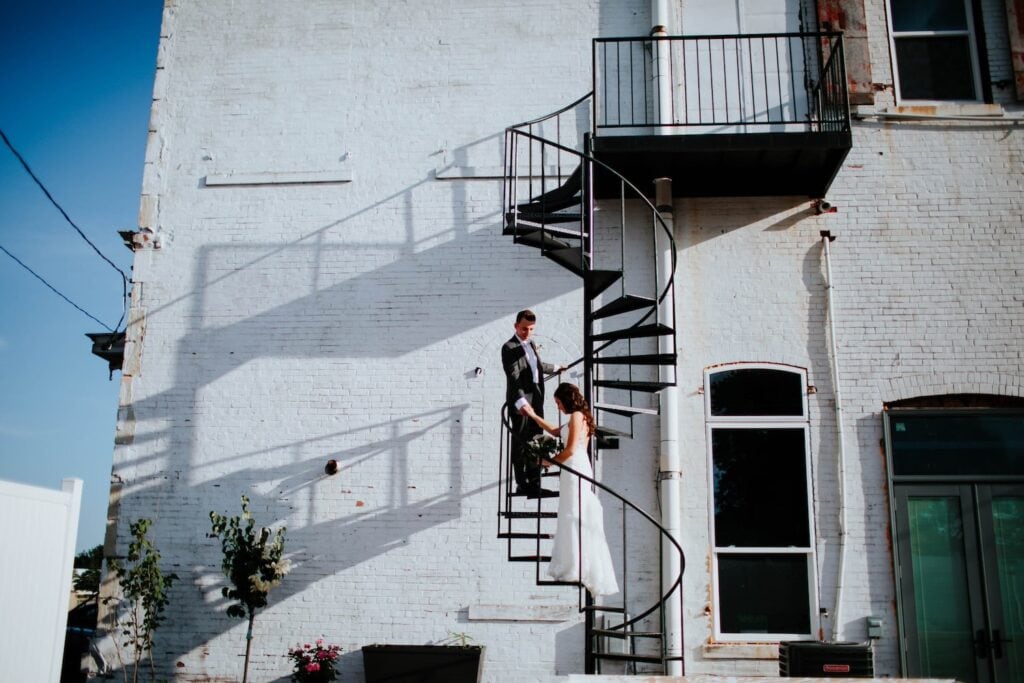 A groom leads his bride up a spiral staircase on the outside of white brick building in the sunshine