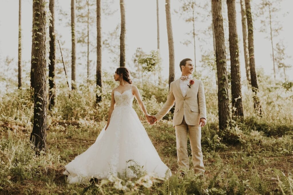 A bride and groom pose in a forest of tall pine trees while holding hands and looking away from each other
