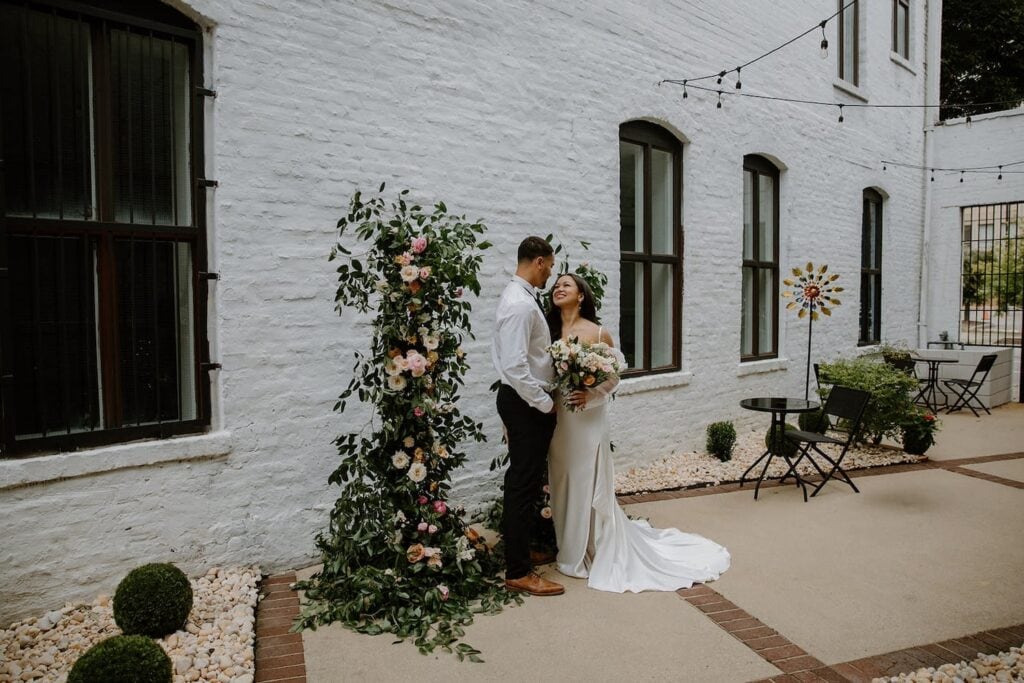 A bride and groom look lovingly at each other in a small alley next to a white brick building with black windows and floral backdrop