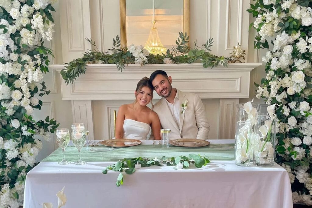 A bride and groom smile at the camera while sitting at a sweetheart table flanked by white floral arrangements