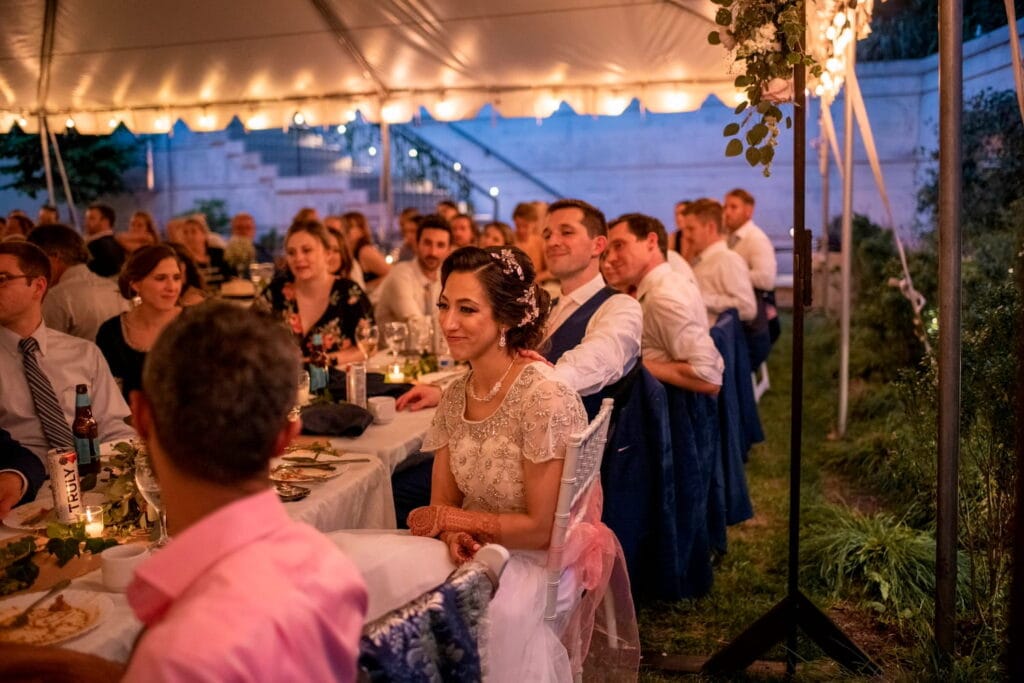 A bride and groom smile while seated under a tent surrounded by friends and family