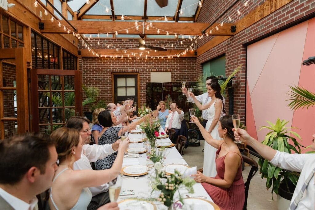 A bride and groom give a toast at a long reception table in an industrial wedding venue