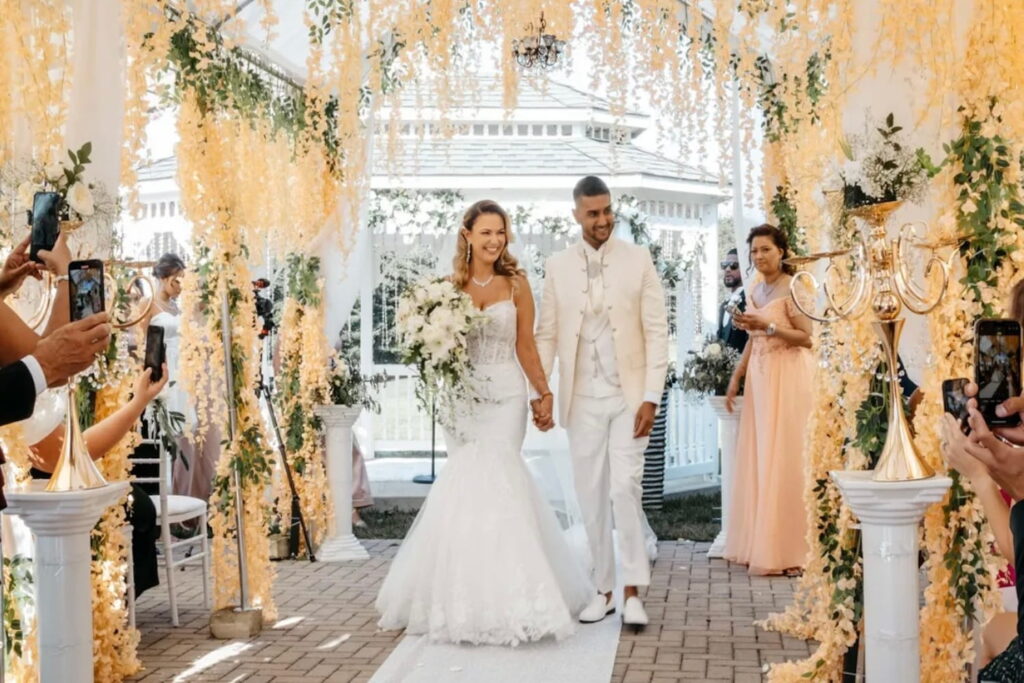 A bride and groom hold hands while walking down an aisle draped in flowers at an outdoor venue