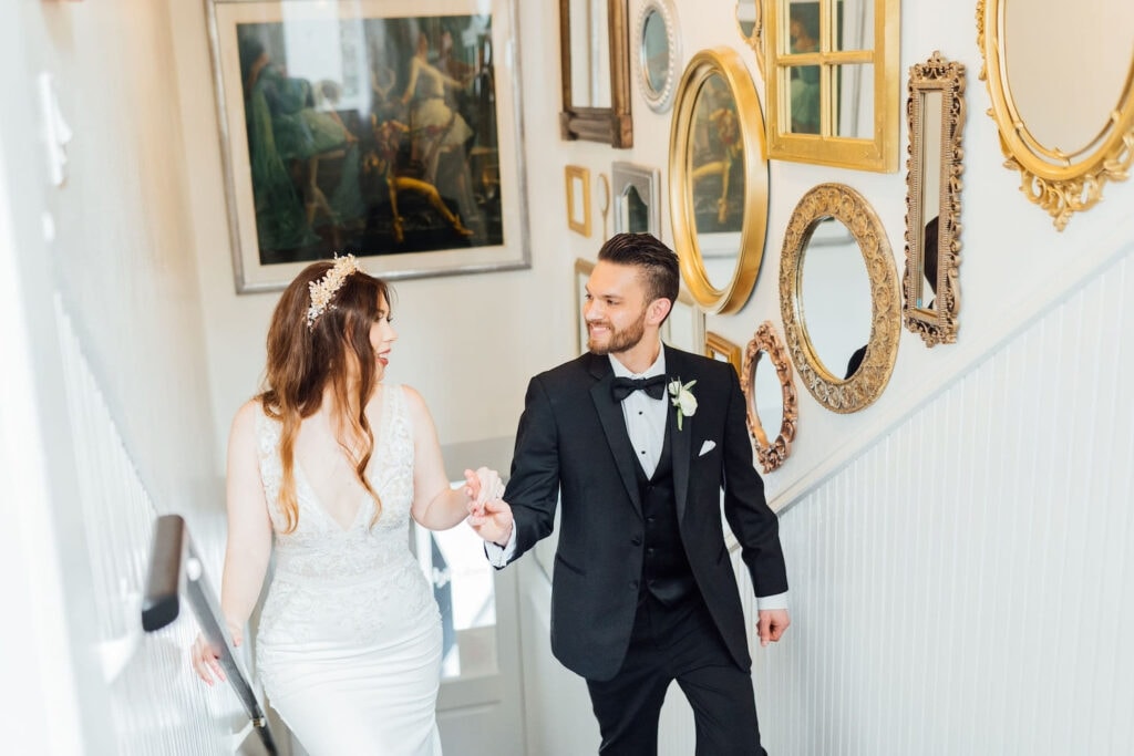 A bride and groom walk up a flight of stairs decorated with gold mirrors hand in hand