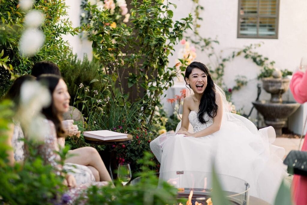 A bride sits in an outdoor reception space laughing with friends and family