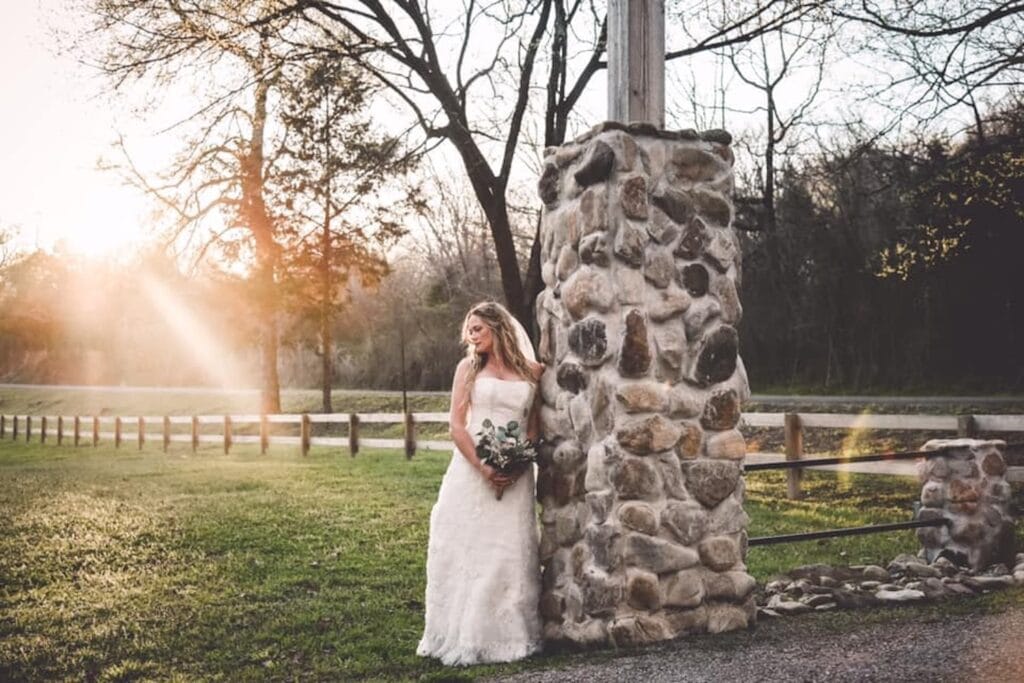 A bride leans against a stone pillar with the sun setting behind her