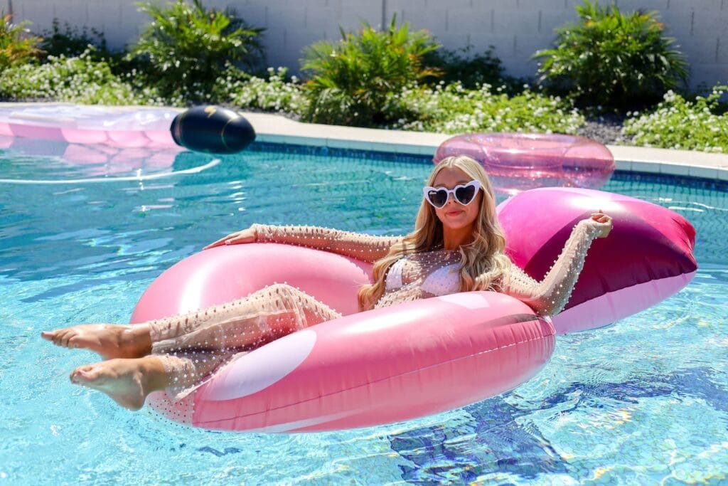 A bride to be in a pool on a pink float and wearing heart-shaped sunglasses smiles at the camera