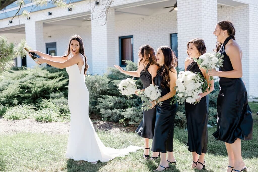 A bride opens a bottle of champagne outside of a brick building as her bridesmaids cheer her on