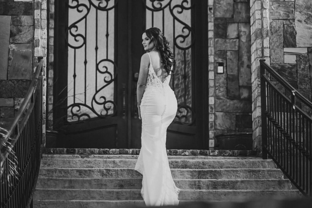 A bride poses in a black and white photo on a stone staircase by an ornately-decorated front door