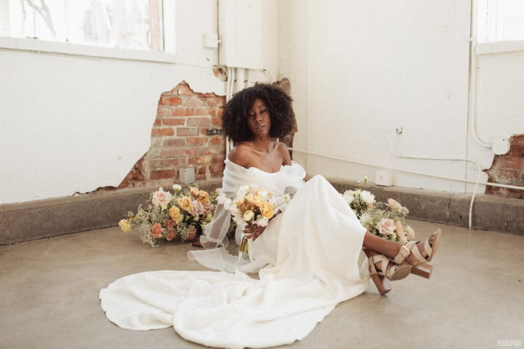 A bride sits on the floor of an industrial space with exposed brick walls, surrounded by floral arrangements