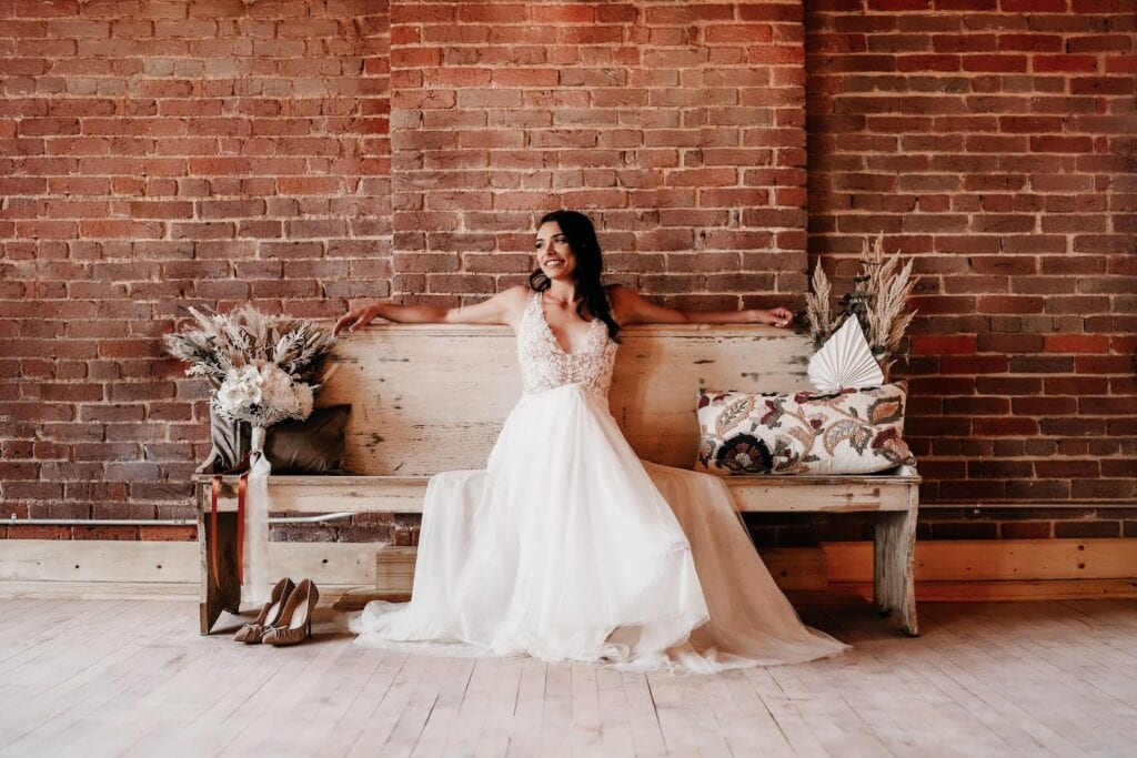 A bride casually sits on a wooden bench decorated with bohemian-style floral arrangements in front of a brick wall with her arms draped across the back of the bench as she smiles and looks off to the side
