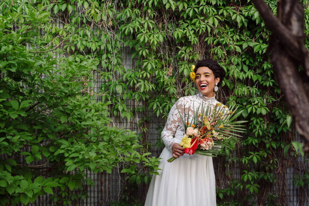 A bride in a long-sleeve lace dress holds a tropical bouquet while standing by wall of greenery