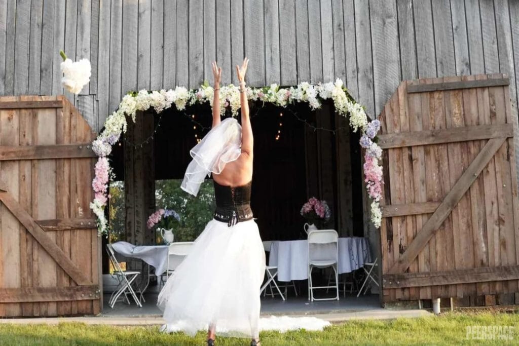 A bride in a leather corset and tulle skirt tosses her bouquet in front of a barn decorated for a reception