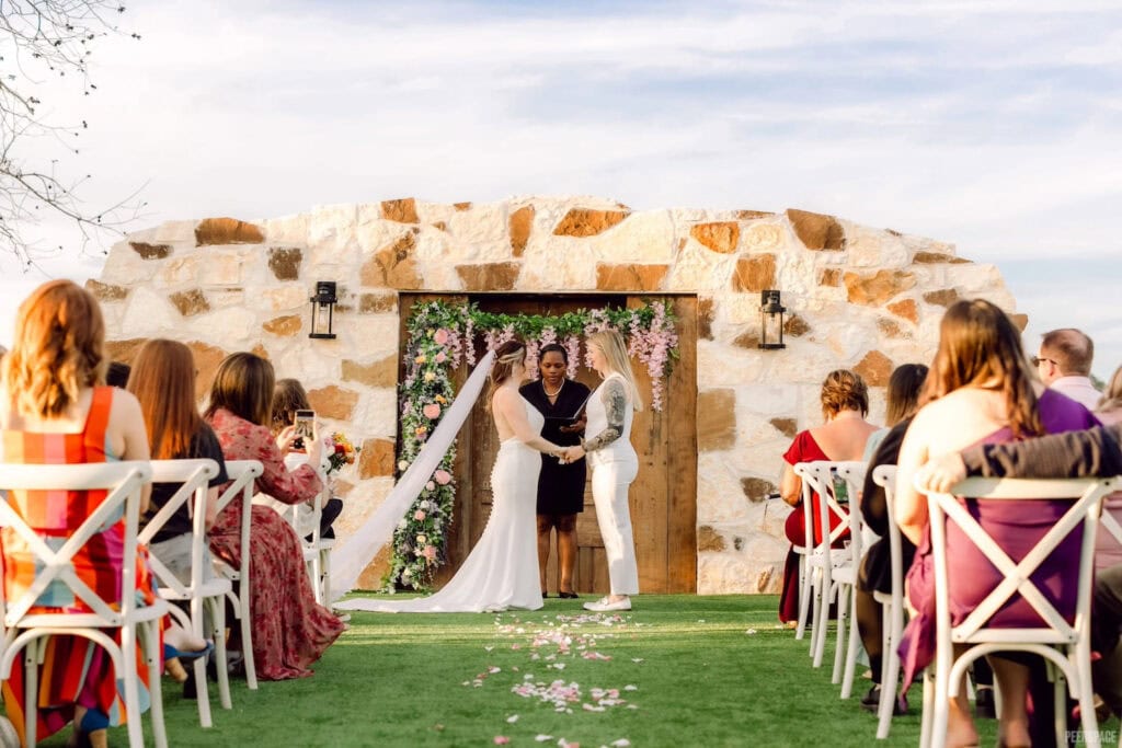 Brides exchange vows outdoors in front of rustic structure decorated with flowers
