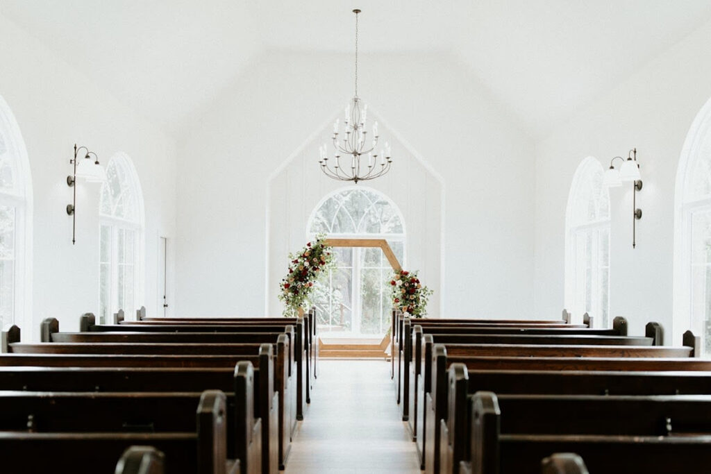 A bright white chapel with dark wood pews is decorated with a wooden hexagon altar covered in autumn-inspired florals