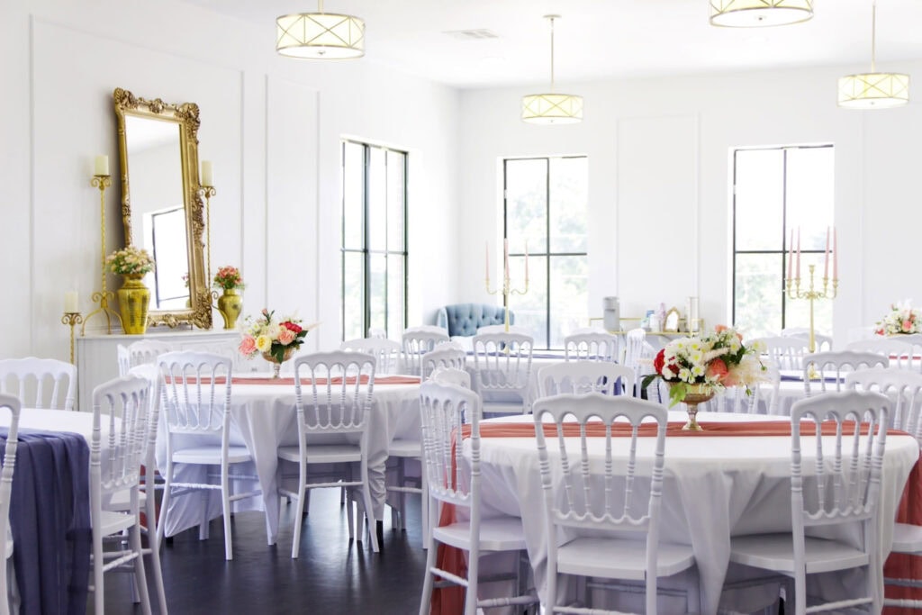 A bright loft space with tall ceilings and windows is set for a reception with all-white tables and chairs and gold, peach and blue accents